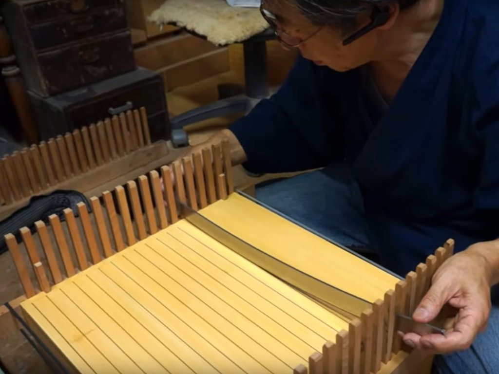 Artisan japonais réalisant l’étape du laquage du shogiban, le plateau du jeu de shogi, à l’aide d’un outil qui ressemble à une lame de katana - source vidéo "Board Making Tachimori by Board Artisan Keiji Miwa”