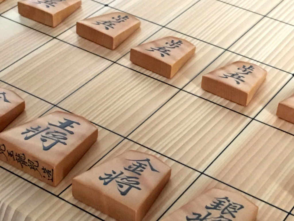 Zoom sur un plateau de shogi sur lequel sont posées des pièces de shogi en bois avec des kanjis noirs calligraphiés par maître Kodama