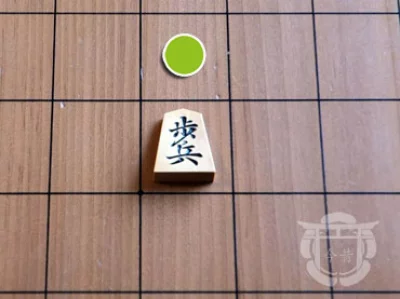 Pièce de shogi en bois sur un plateau en bois, ici le pion, avec son déplacement indiqué par un symbole vert